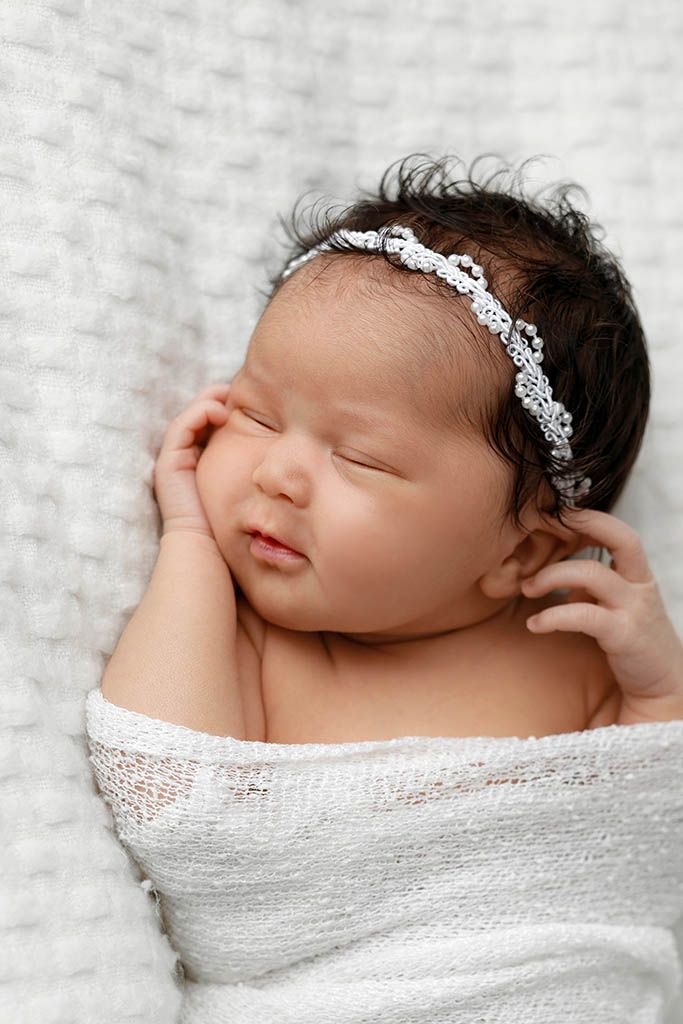 Peaceful sleeping newborn baby girl swaddled in white wrap wearing delicate rhinestone headband.