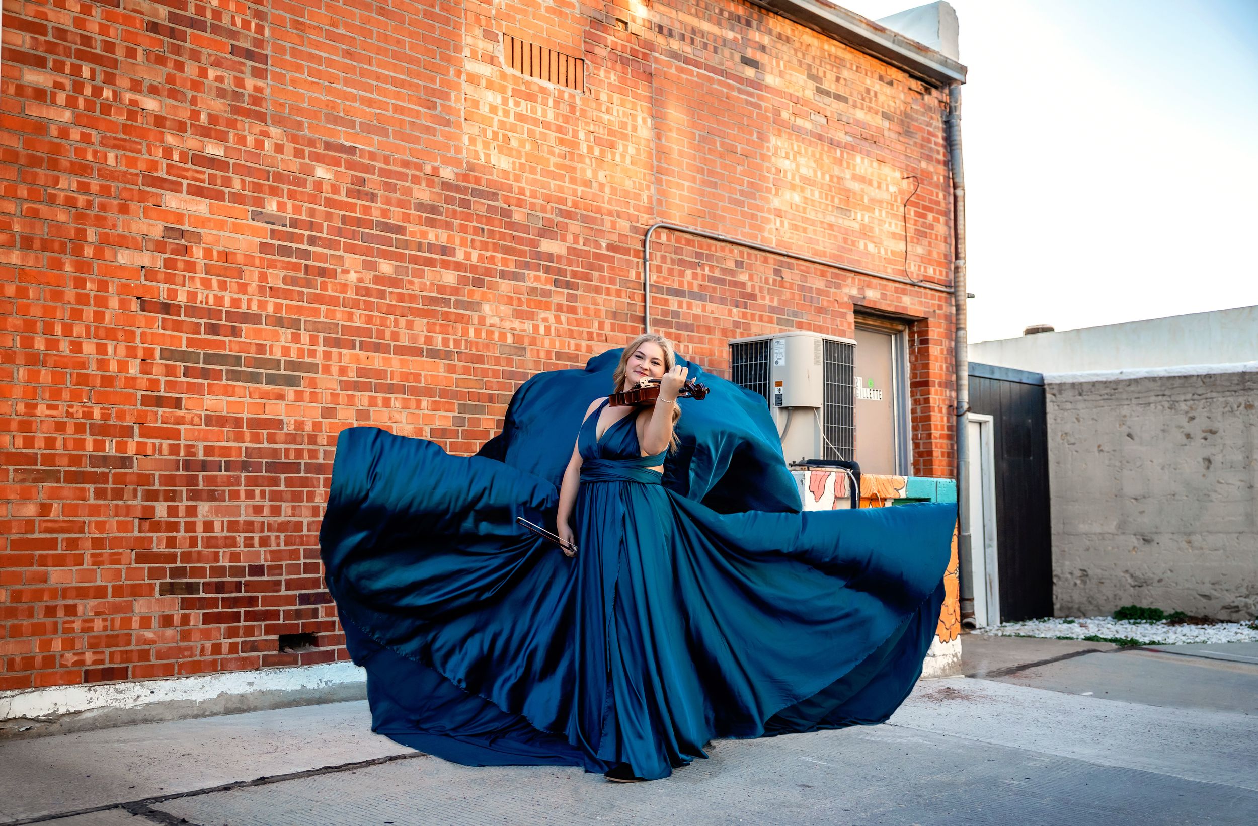Woman in flowing navy blue gown poses dramatically outdoors against a red brick wall.