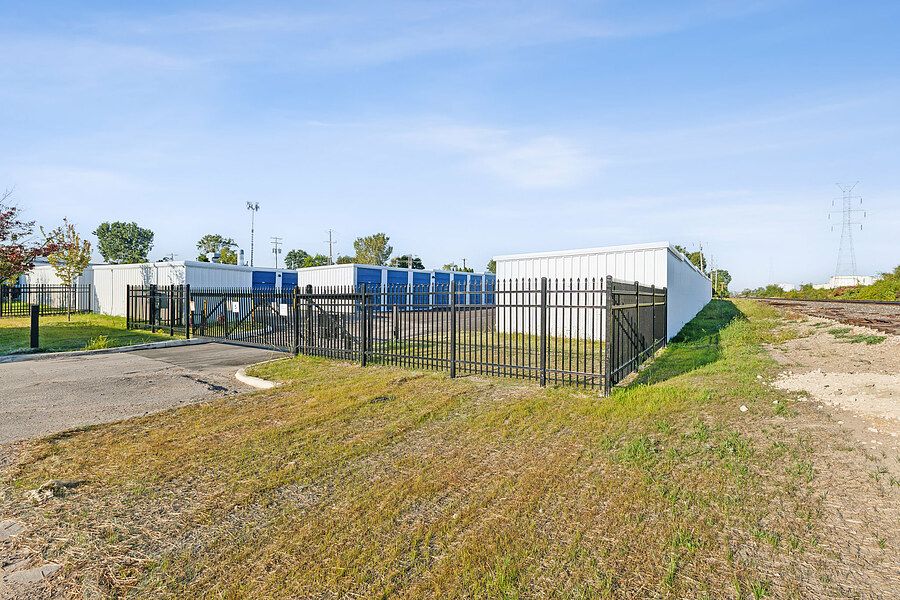 Outdoor self-storage facility with blue units behind black iron security fence on green lot.
