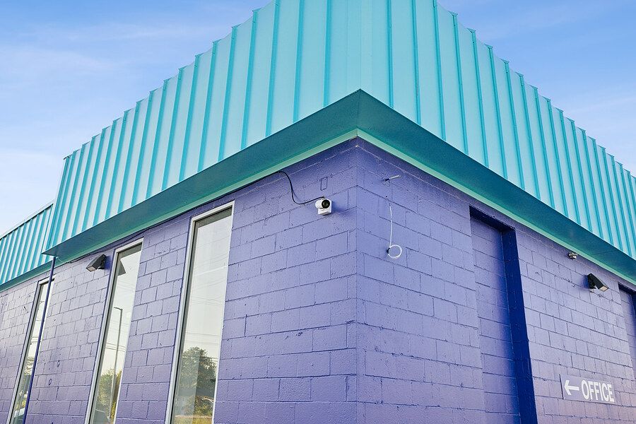 Purple brick storage facility with teal metal roof panel siding and security camera mounted on exterior wall.