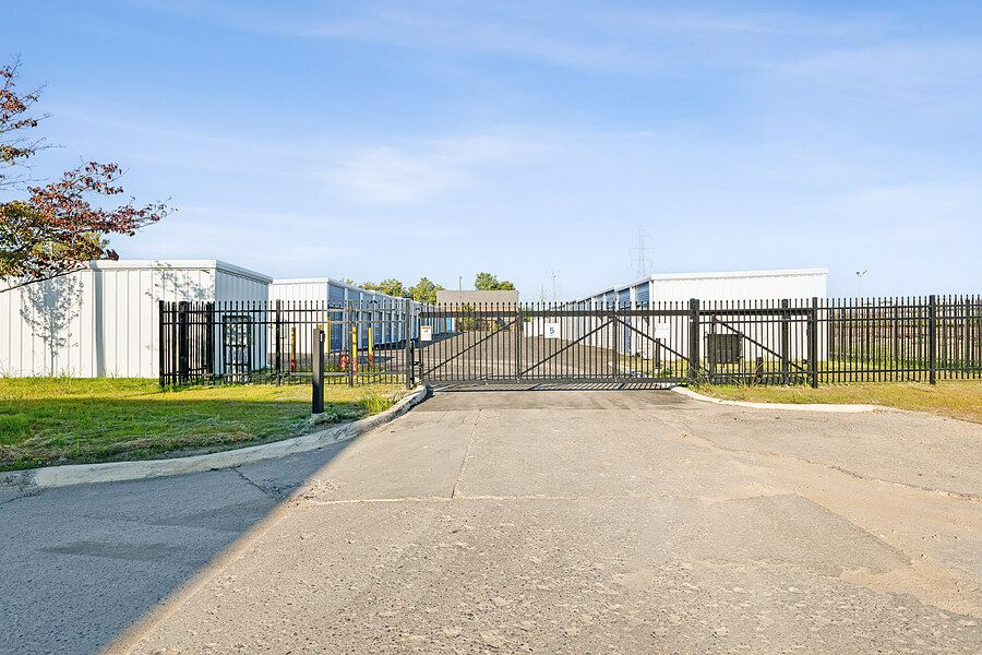 Black iron security gate entrance to a fenced storage facility with white buildings under a clear blue sky.