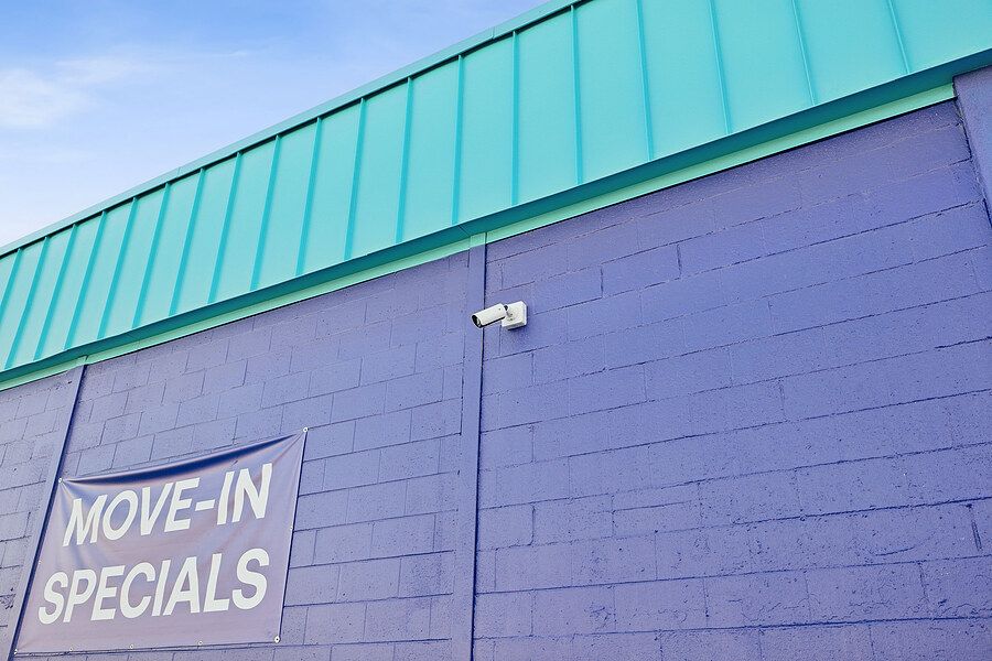 Purple brick building with teal metal roof displaying Move-In Specials sign and security camera under blue sky.