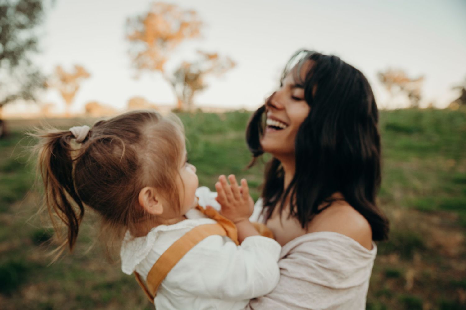 A laughing mother holds her happy toddler girl in her arms in a sunlit green field.