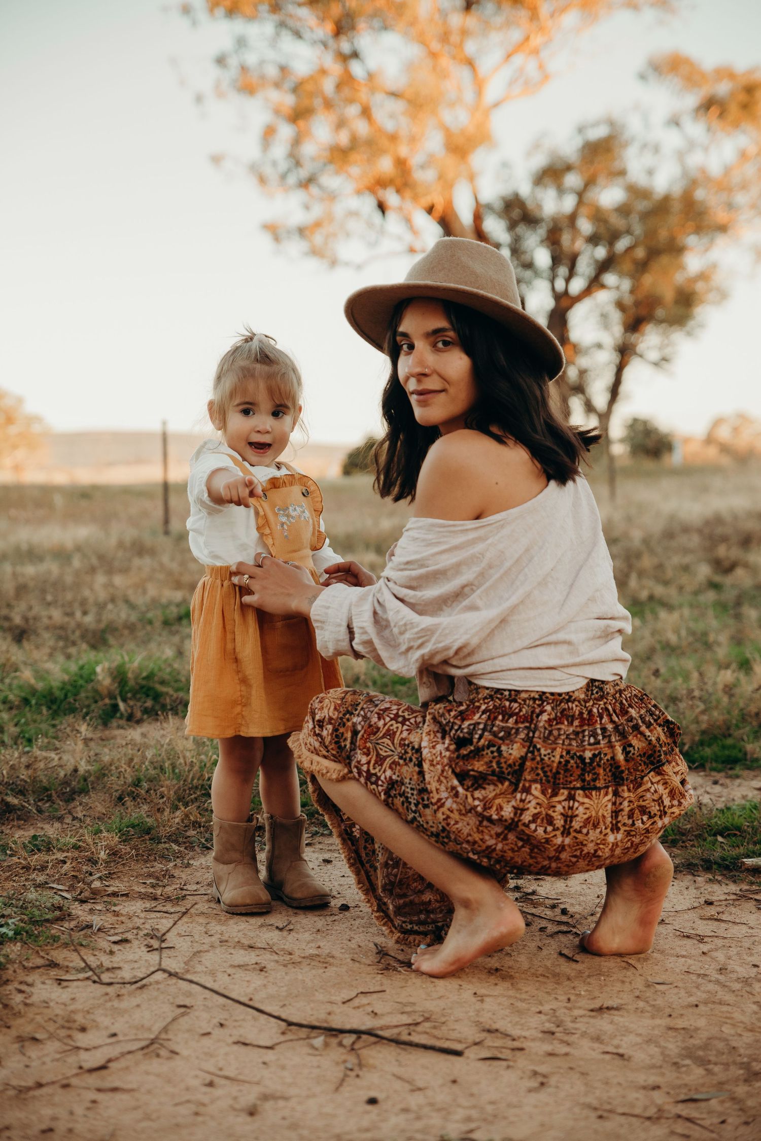 A barefoot mother in a wide-brim hat crouches beside her toddler daughter on a dusty rural path.