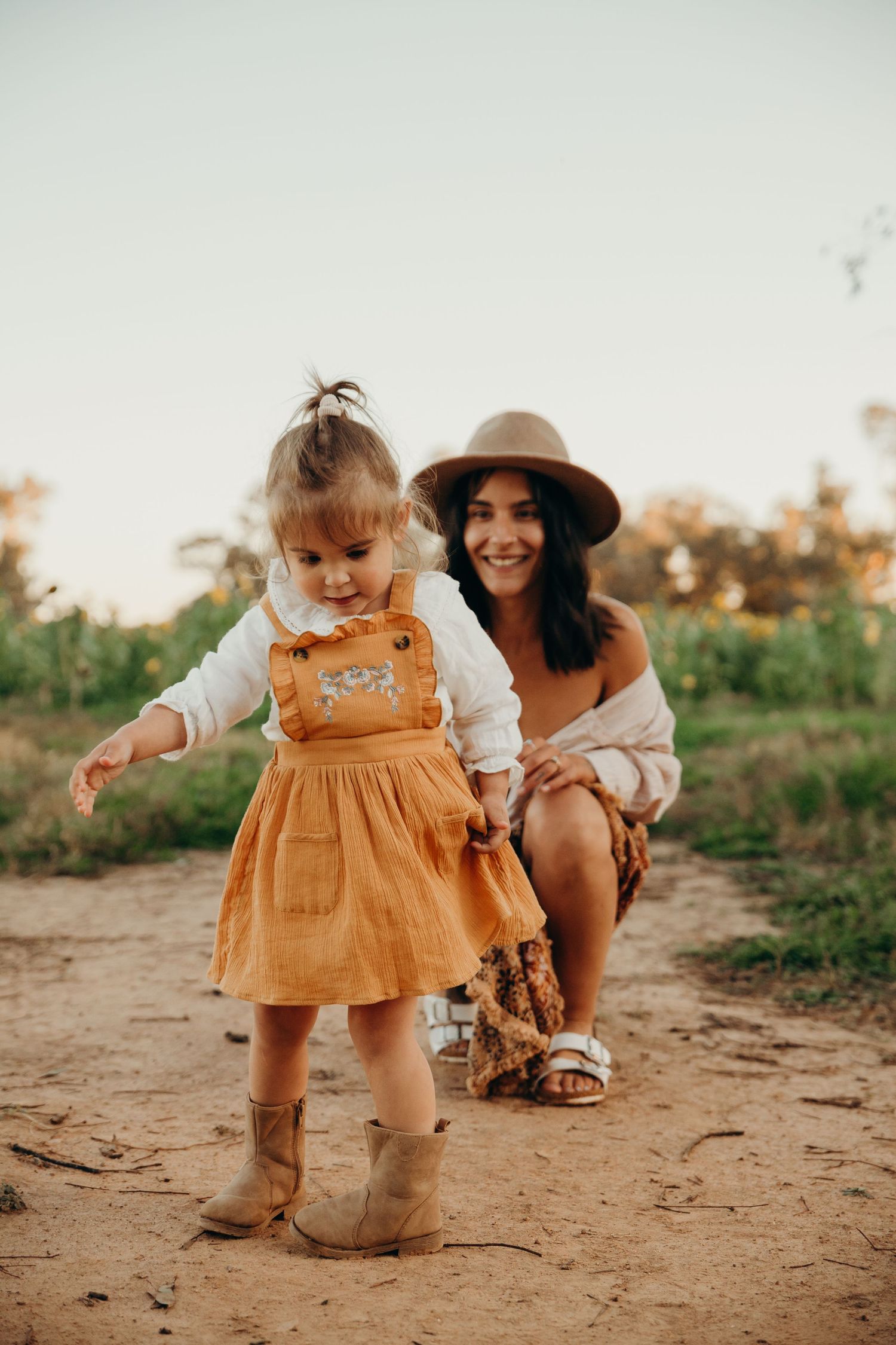 Mother in boho hat crouches beside curious toddler girl in mustard overalls on a sunny dirt path outdoors.