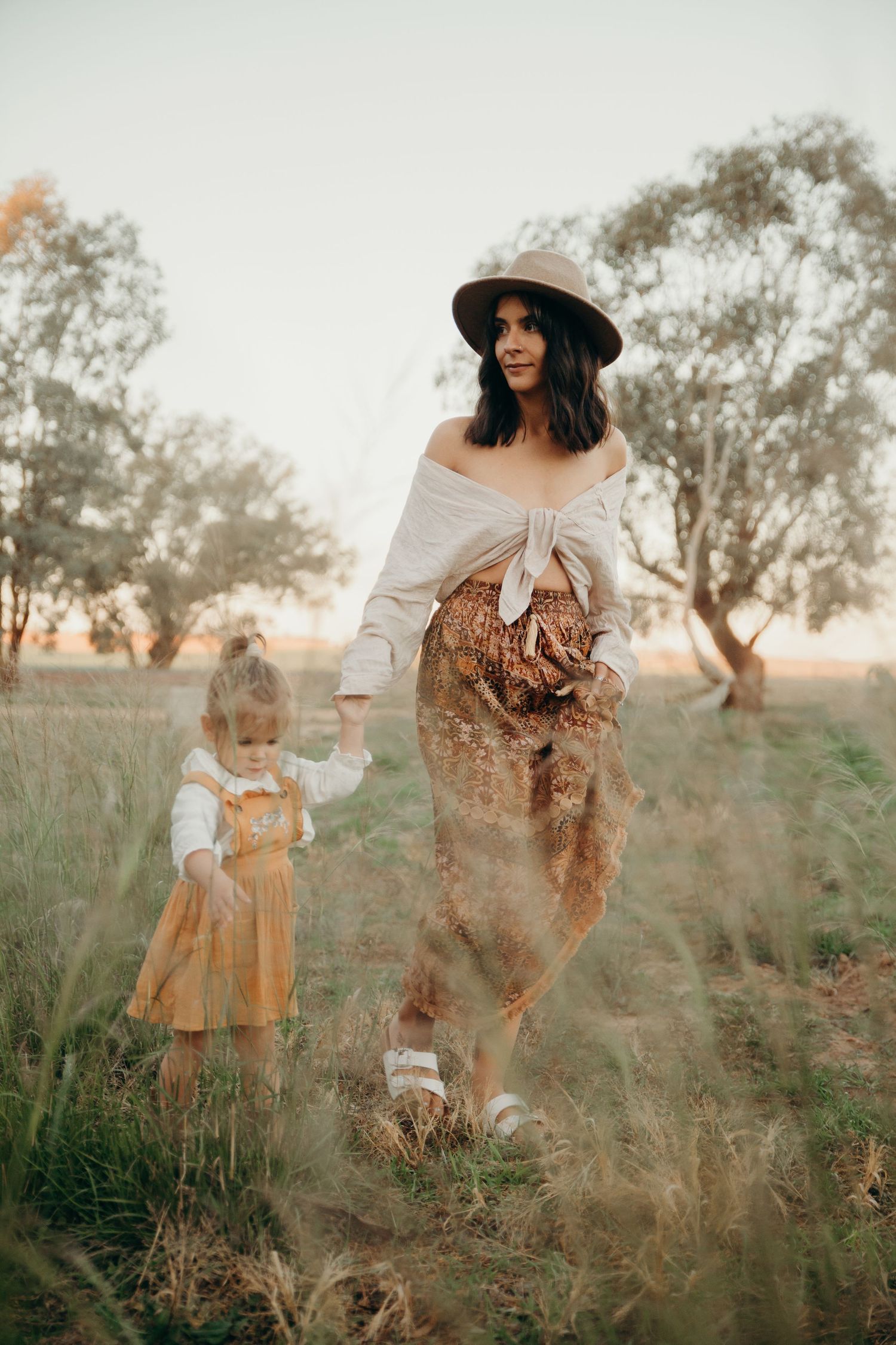 Boho mother in hat walks hand in hand with toddler daughter through golden grass field at sunset.
