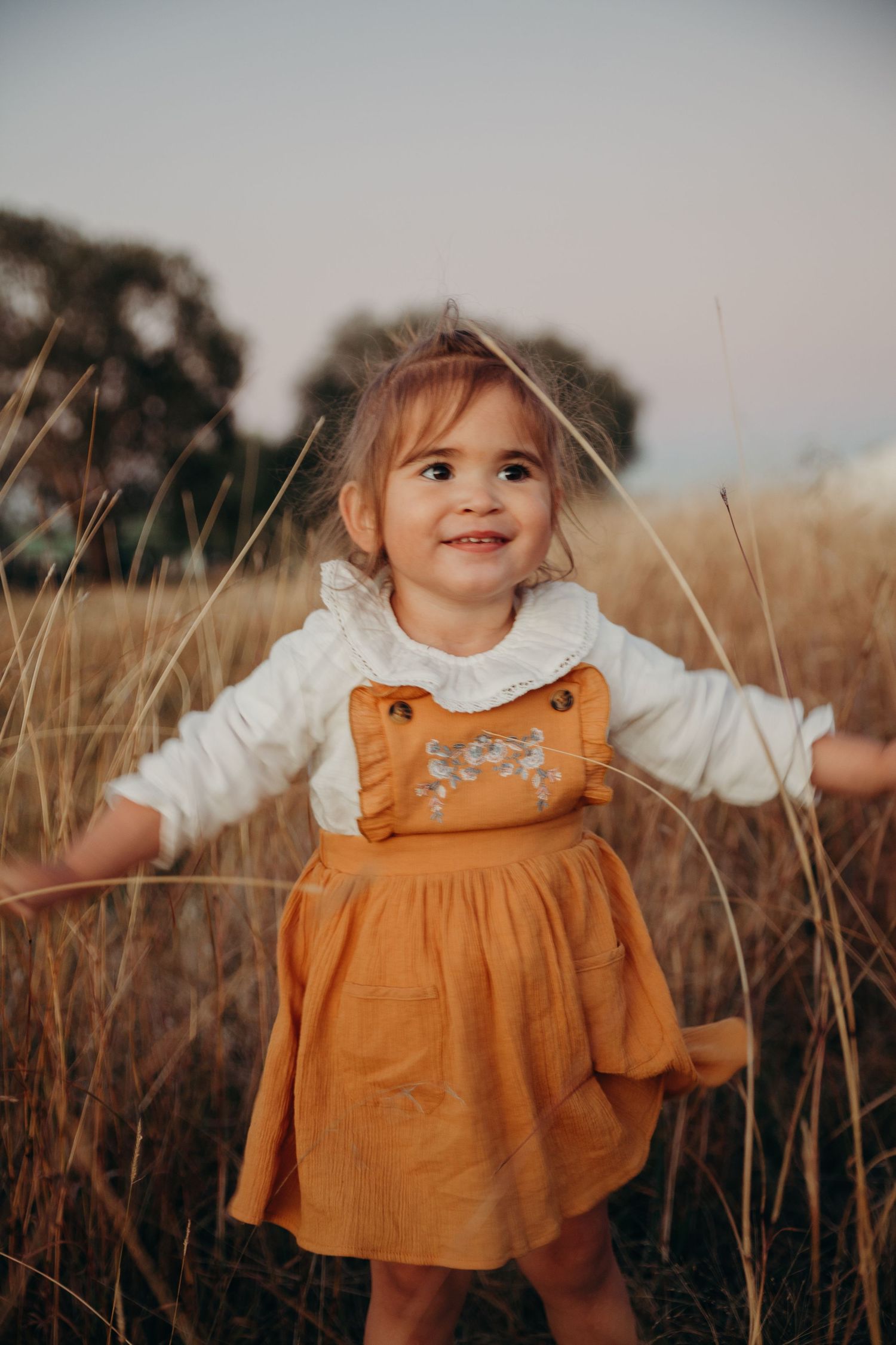 Smiling toddler girl in mustard dress and white top playing in tall golden grass field.