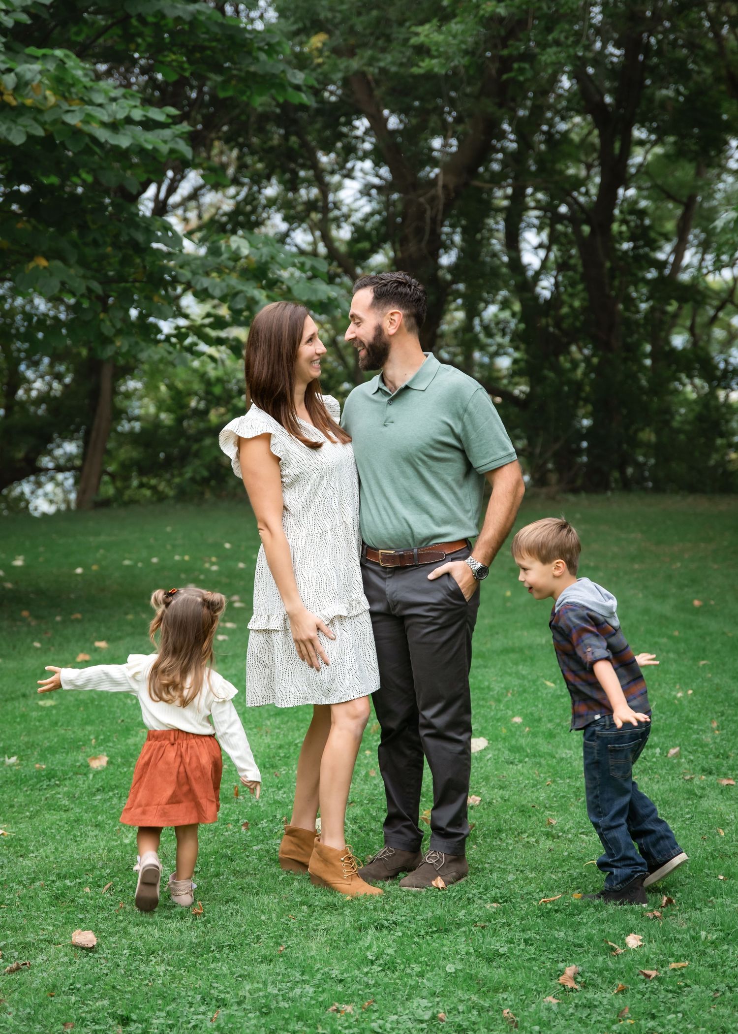 Family of four posing outdoors on green grass with tall trees, parents smiling at each other while two young children play.