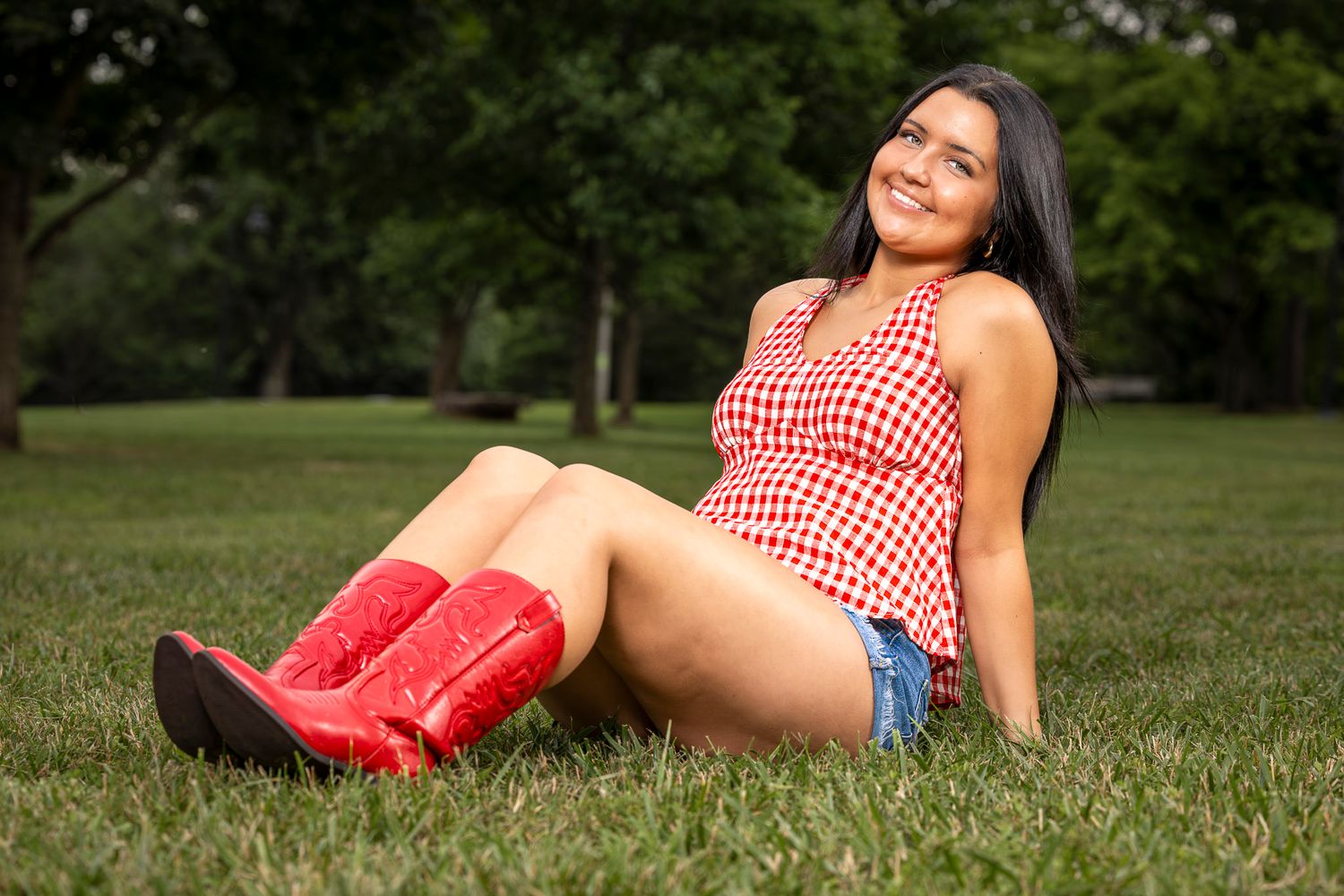 A smiling woman in a red checkered top, denim shorts, and red cowboy boots sits on green grass in a park.