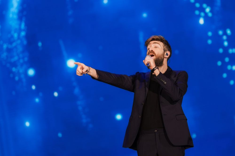 Male performer in black suit singing on stage with a blue light background.