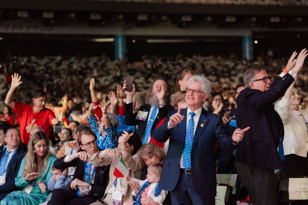 Enthusiastic crowd with raised hands at a large conference, led by a man speaking into a microphone.