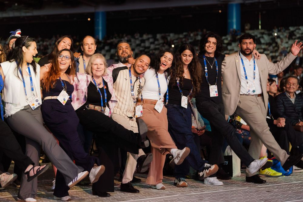 A diverse group of young professionals pose playfully at an international conference event wearing lanyards.