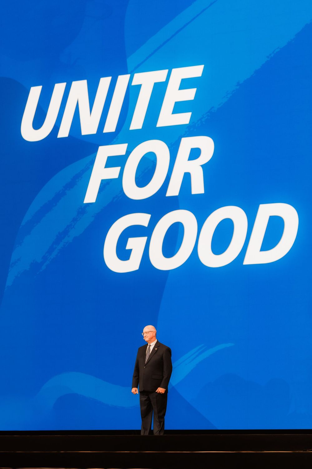 Man standing on stage beneath large blue Unite For Good Rotary International sign.