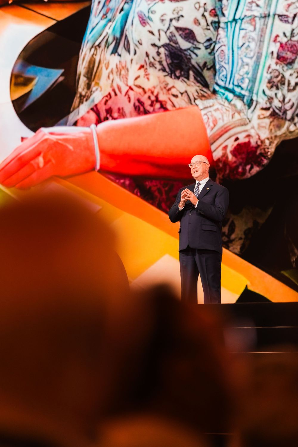 A man in a dark suit speaks on stage before a large colorful projected backdrop at a corporate event.