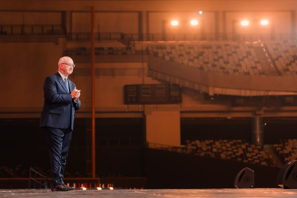 Man in blue suit stands alone on large stage in empty auditorium under warm stage lighting.