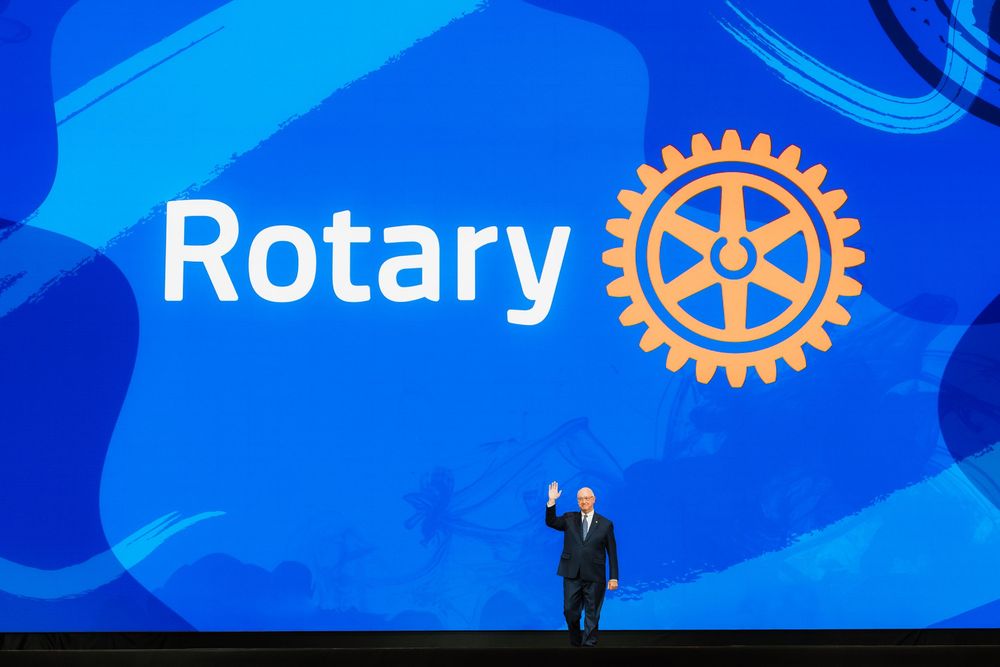 Speaker on stage at Rotary International event with large blue backdrop and Rotary logo.