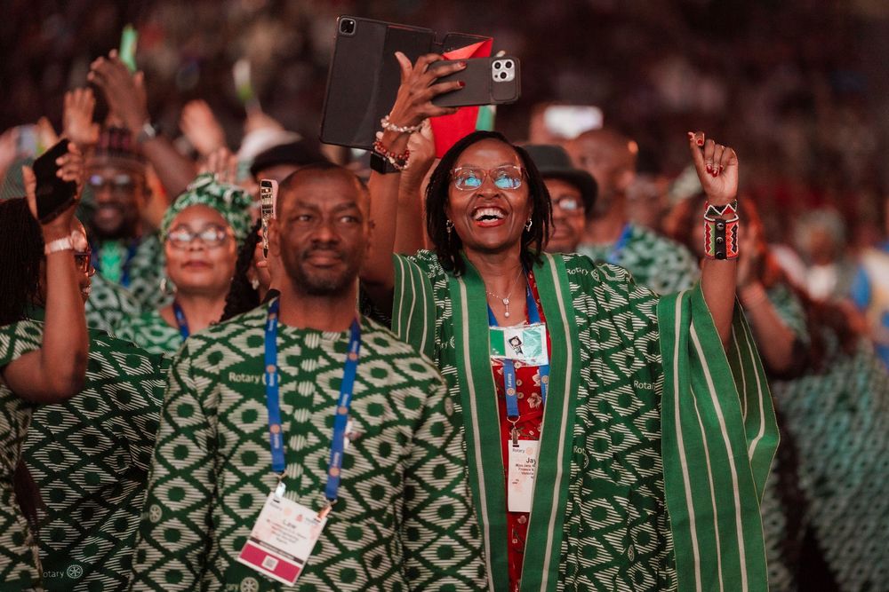 Delegates in green traditional African attire celebrate at a ceremony, with a woman taking a selfie on a tablet.