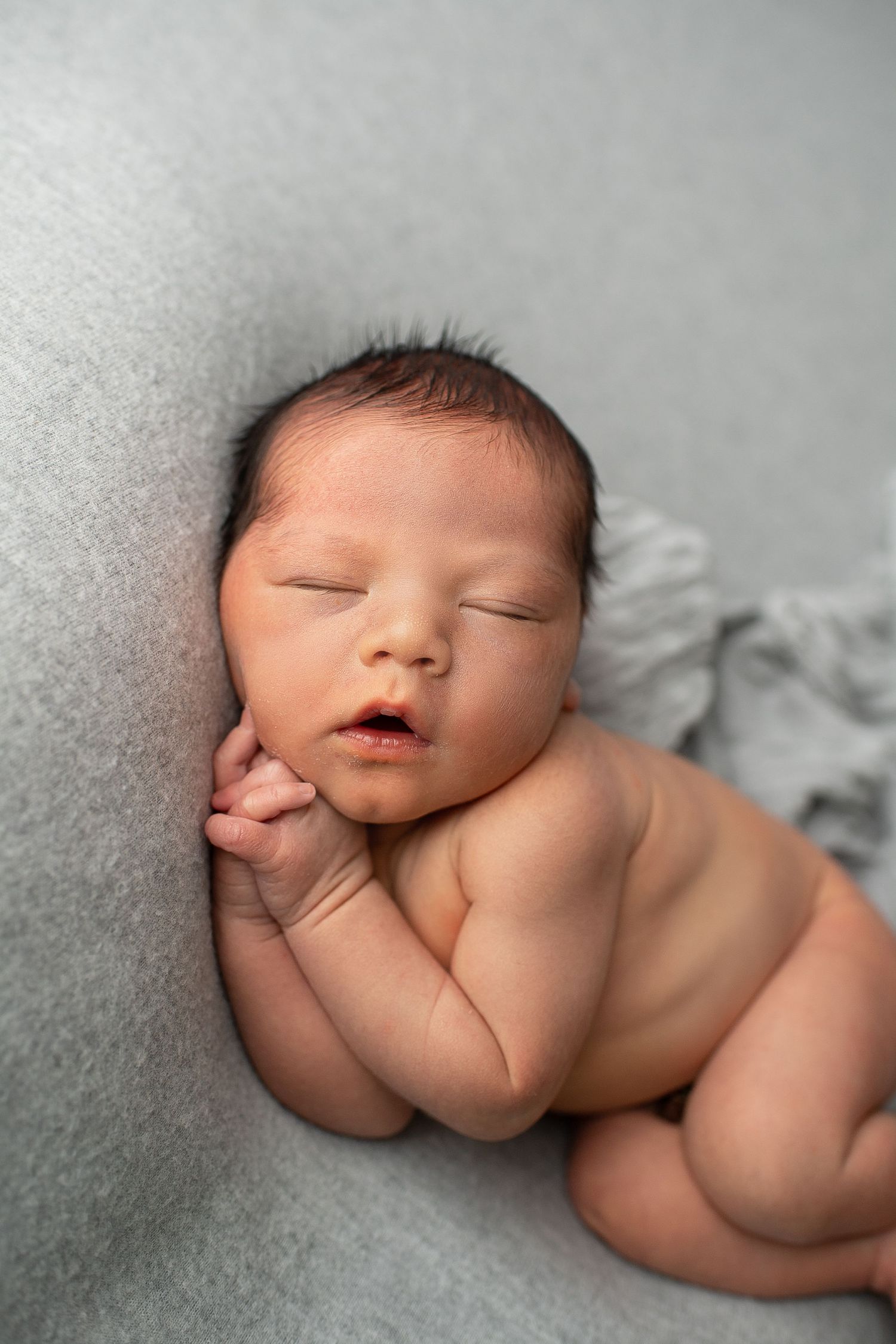 Peaceful newborn baby sleeping on a gray blanket with mouth slightly open.