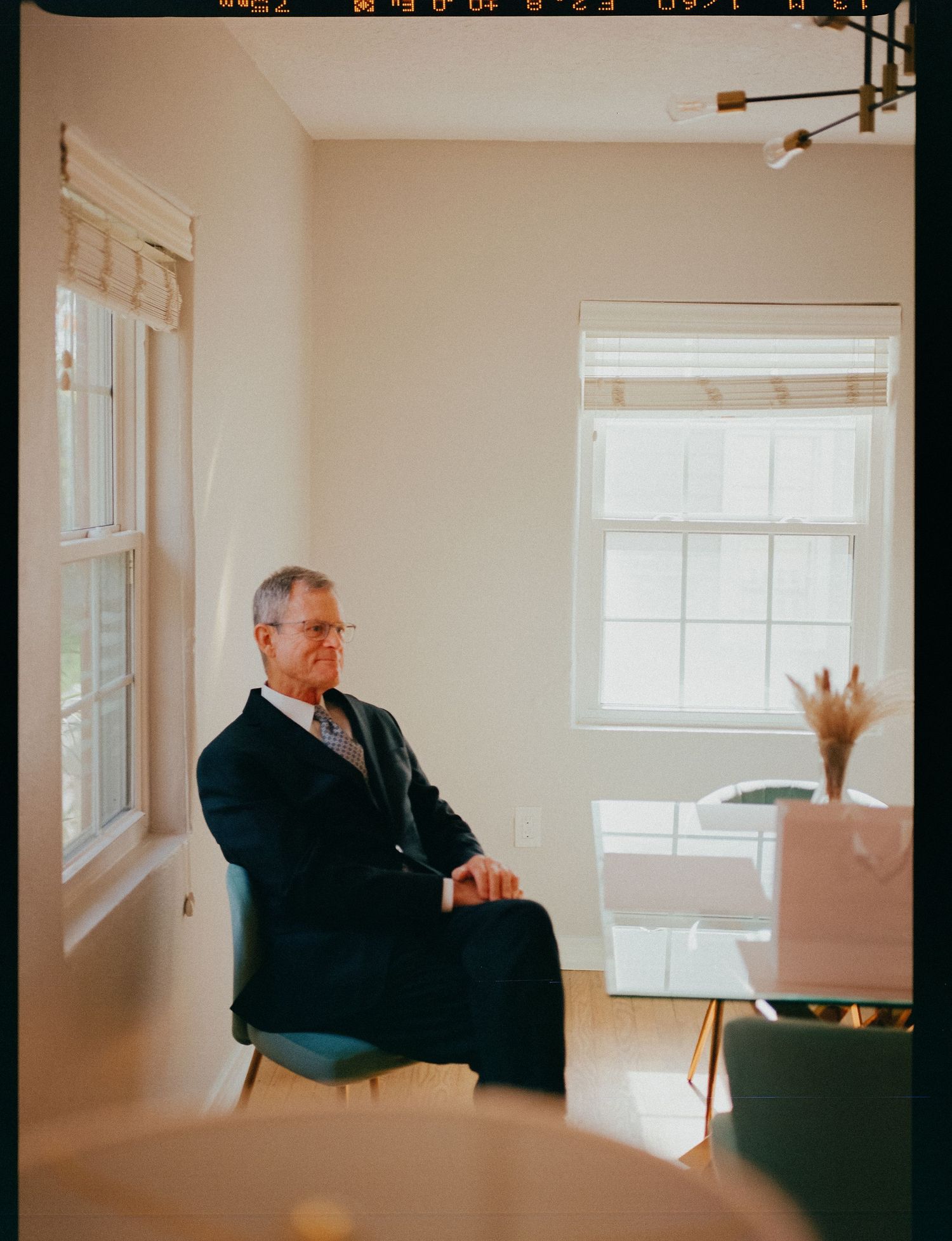 Older man in navy suit sitting pensively in a bright, modern room with large windows and hardwood floors.