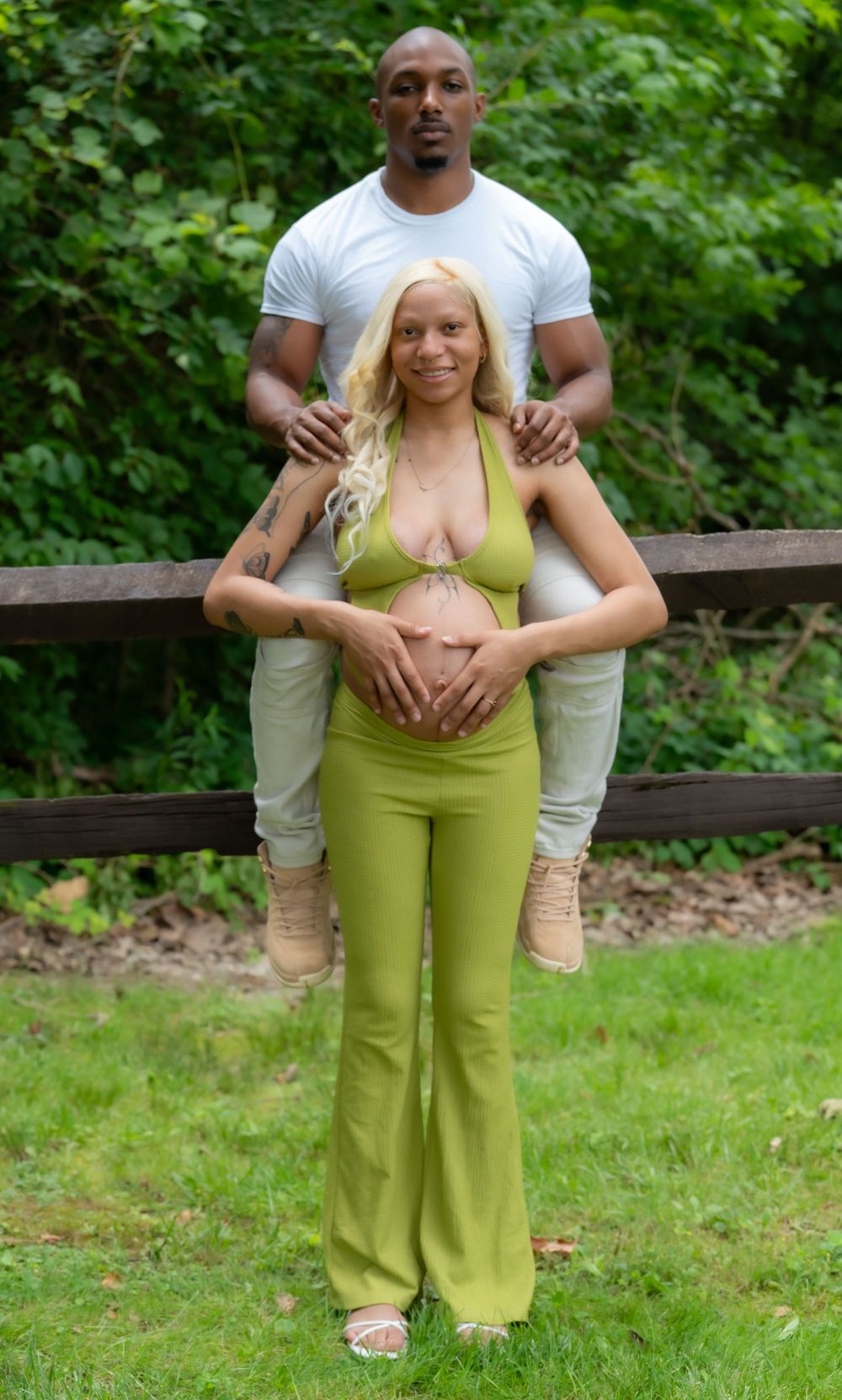 A couple poses outdoors, man in white tee behind pregnant woman in green crop top and flared pants, hands on her belly.
