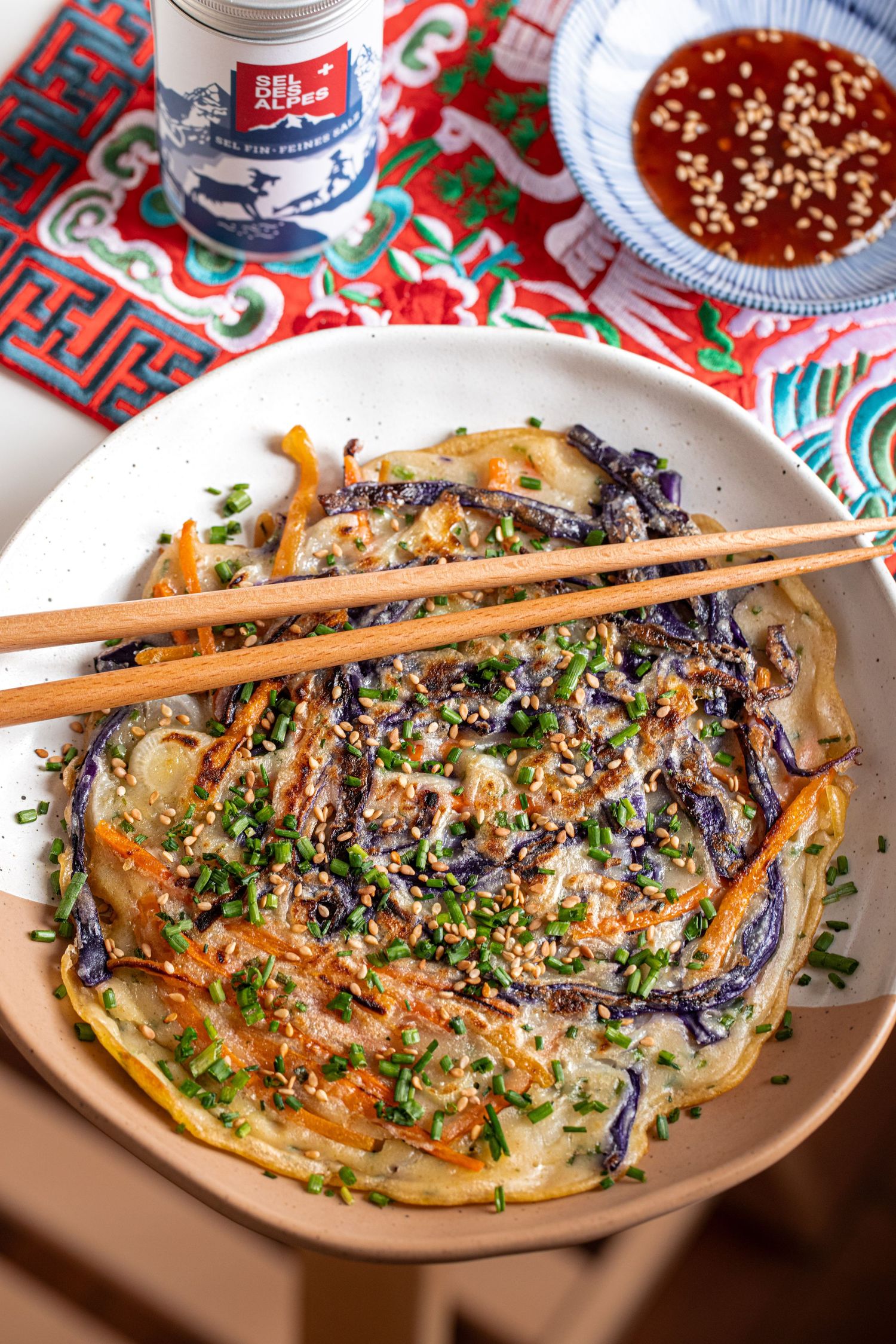 Savory Korean pajeon pancake with purple cabbage, carrots, and chives, topped with sesame seeds, served with dipping sauce.