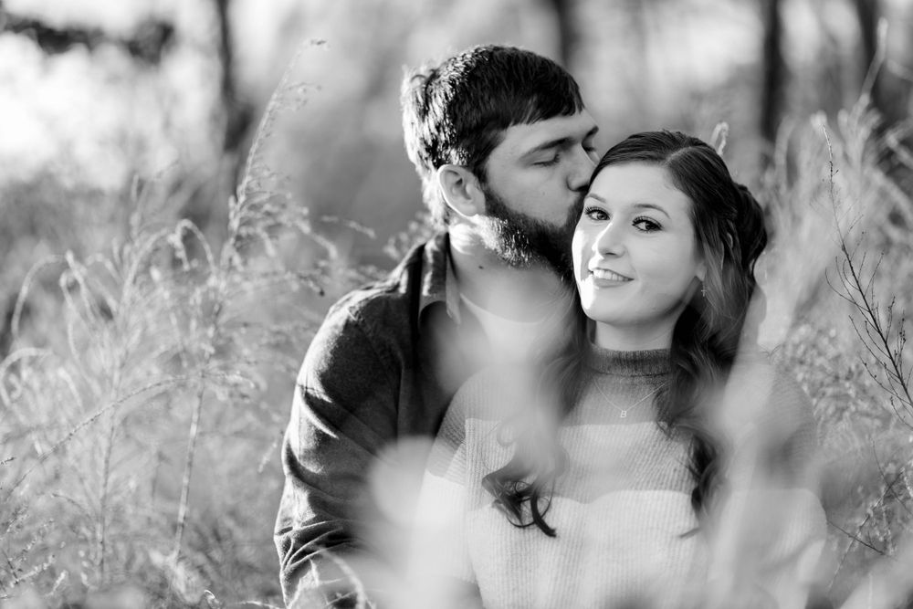 Black-and-white photo of a man kissing his partner's temple in a grassy field during a couples session in Mountain Home, Arkansas