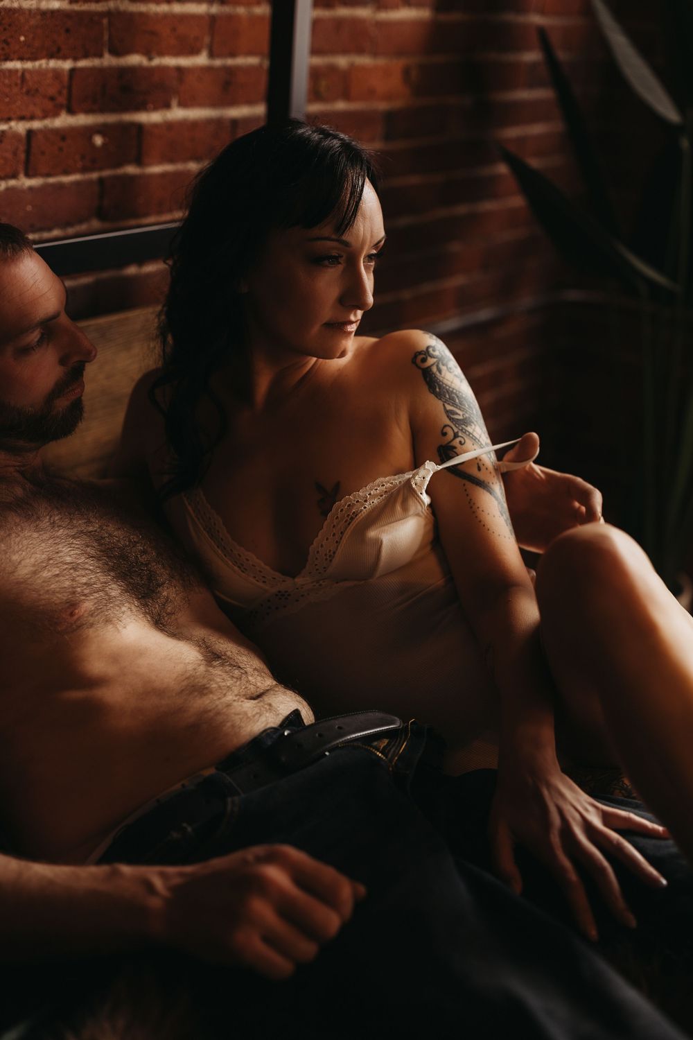 Intimate couple in moody brick loft, tattooed woman in lingerie resting against shirtless man in dim light.