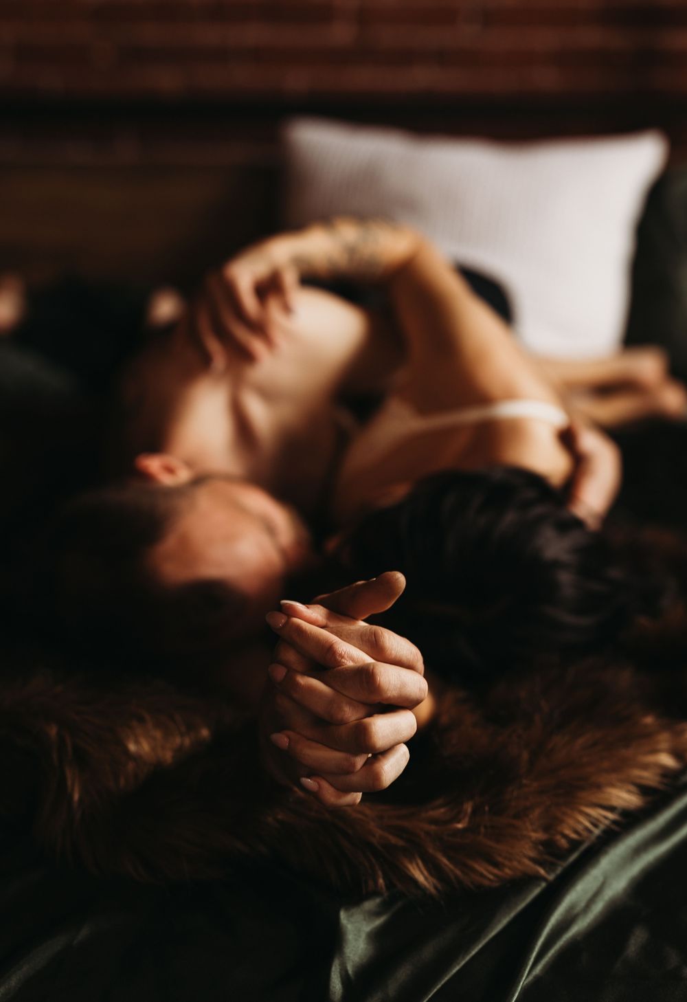 Two people lying on a fur blanket holding hands in a romantic and intimate bedroom setting.