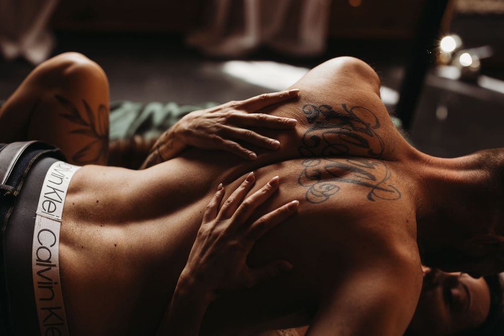 Tattooed person lying down wearing Calvin Klein underwear, with hands resting on their back in moody lighting.