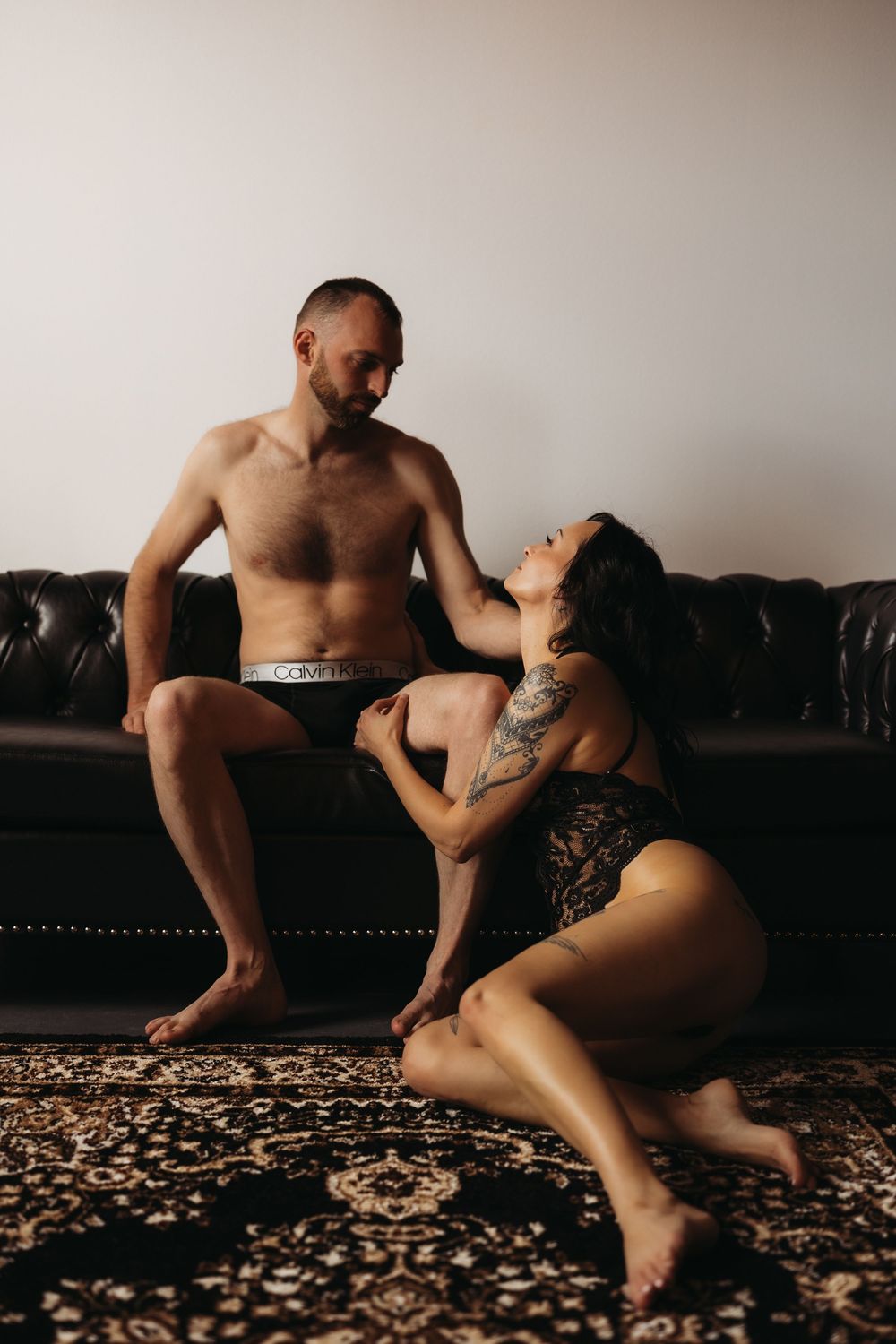 Man and woman in intimate boudoir session posing on leather couch in elegant black and white lingerie.