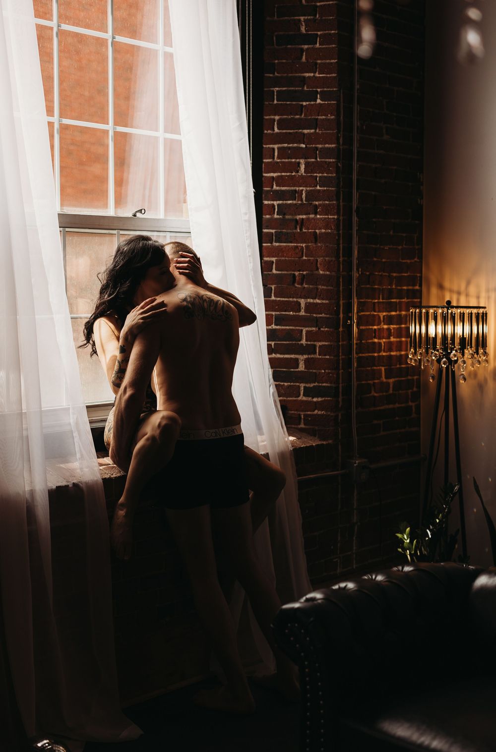 Two silhouetted figures embrace intimately by a sunlit window with sheer curtains and exposed brick wall.