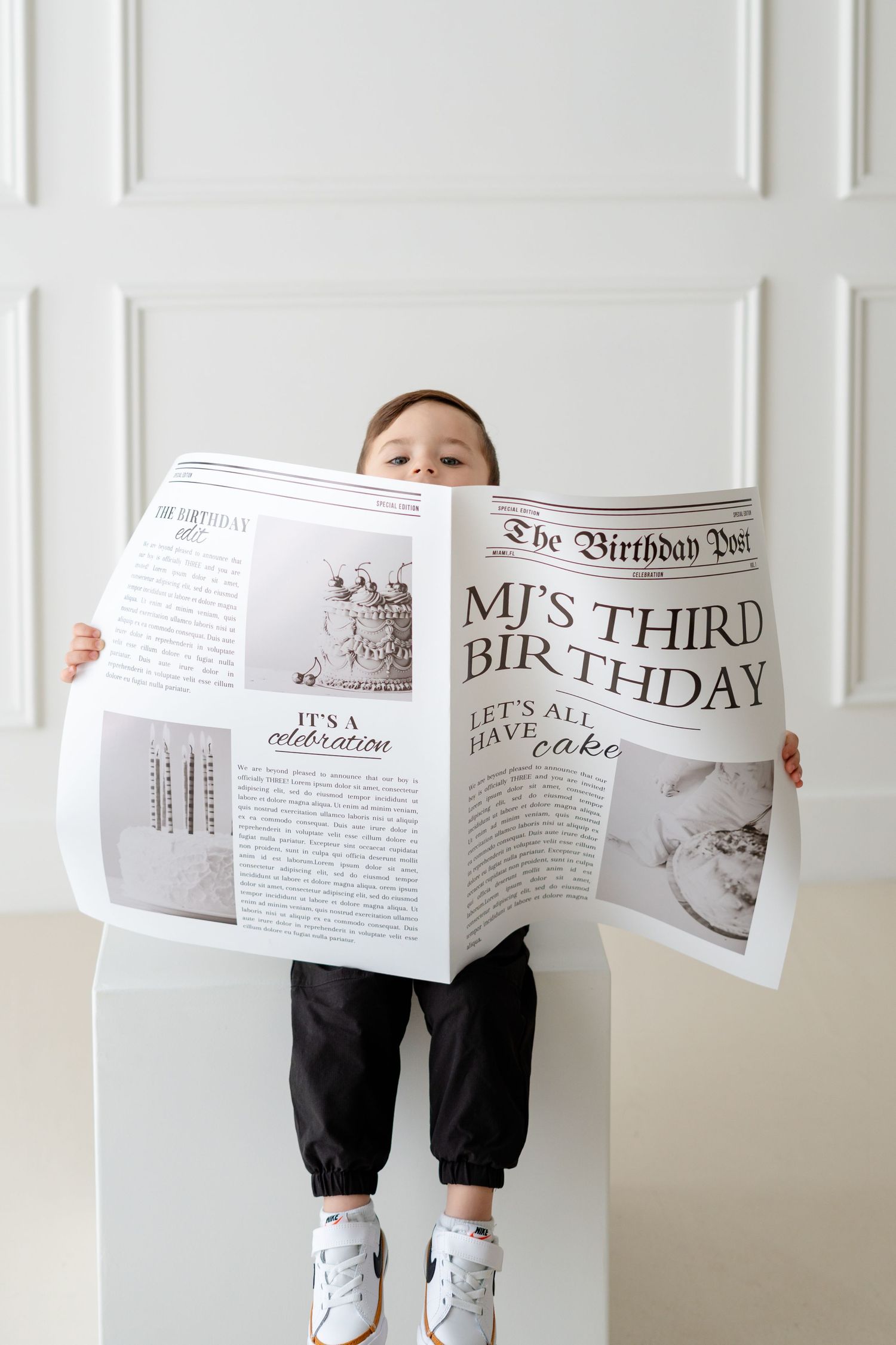 Young child holding a large Birthday Post newspaper celebrating MJ's third birthday against a white wall.