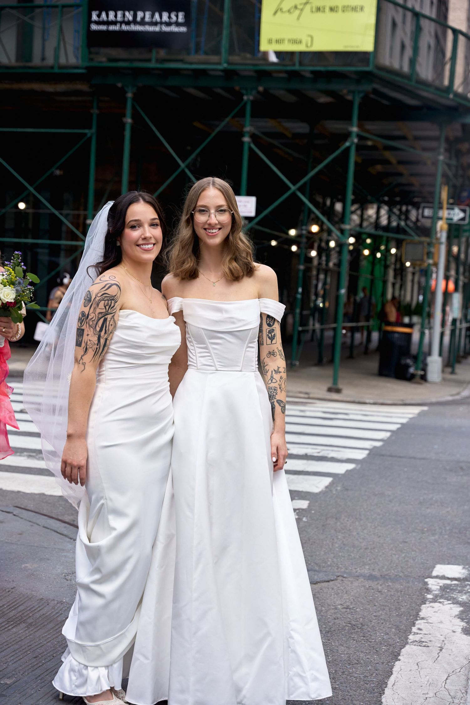 Two brides in white gowns with tattoos pose on a NYC street corner, one wearing a veil holding flowers.