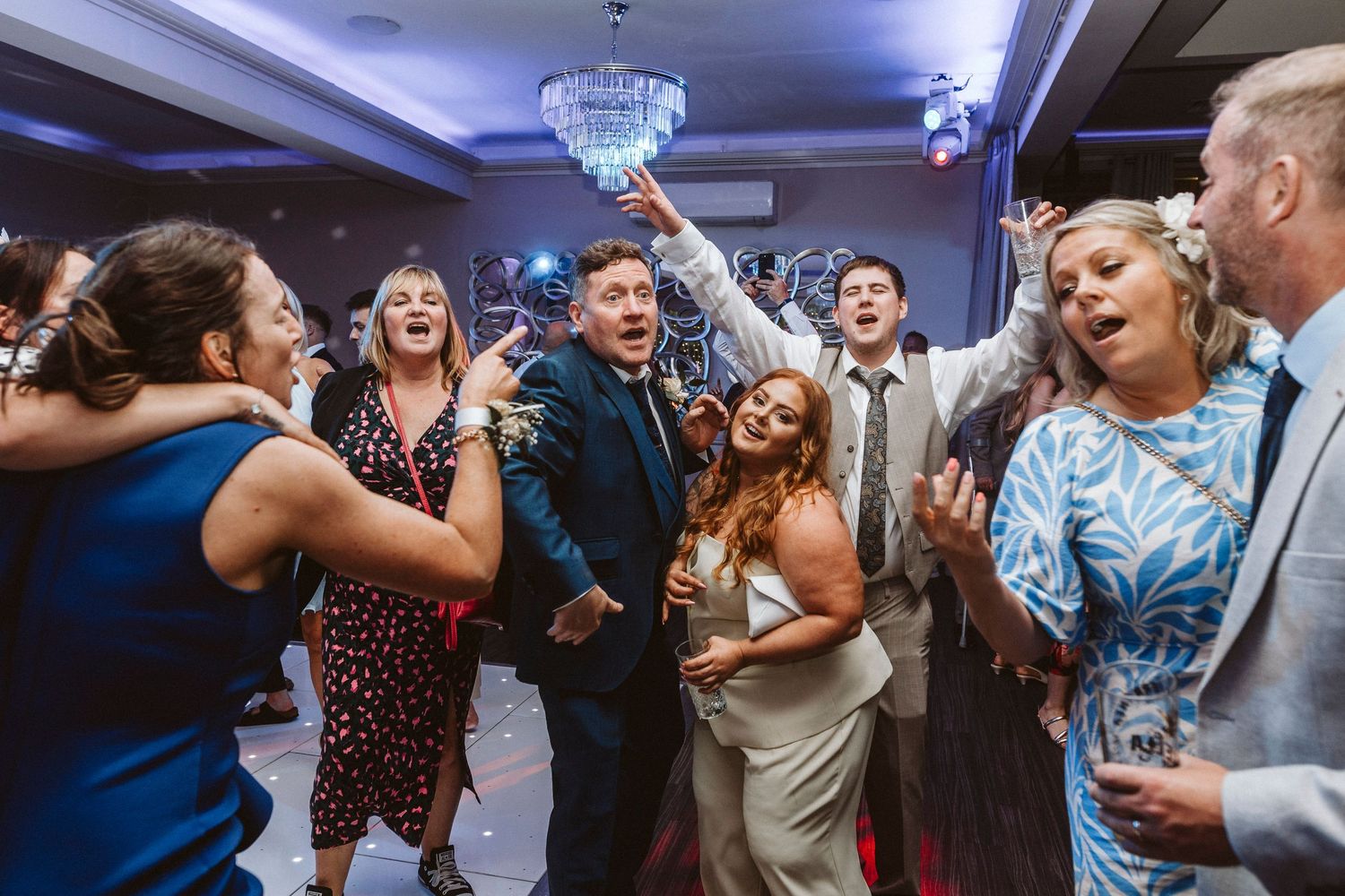 Joyful wedding guests dancing and celebrating together in an elegant venue with chandelier lighting.