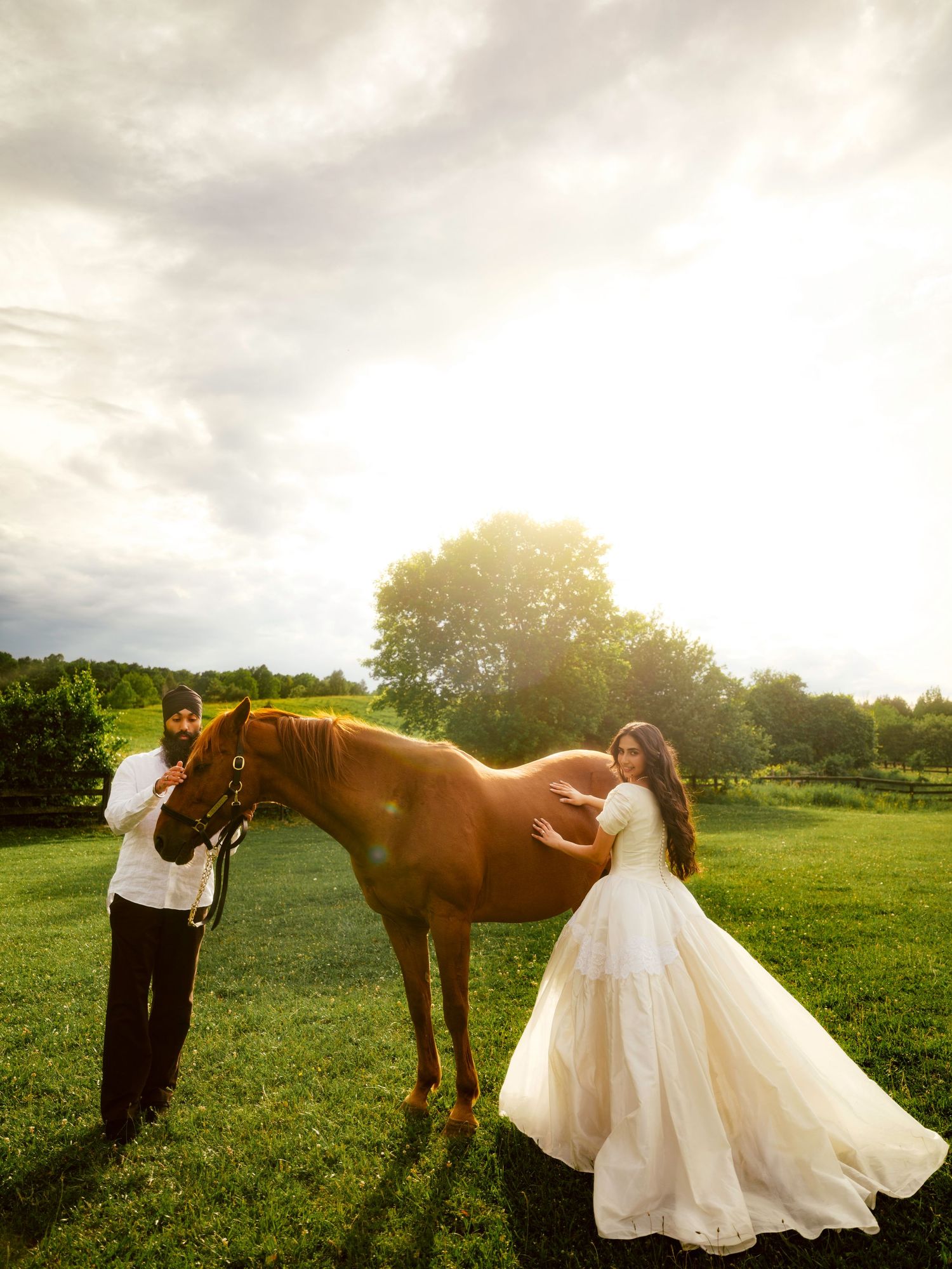 Couple in white attire poses with a chestnut horse in a sunlit green field during a romantic outdoor engagement session.