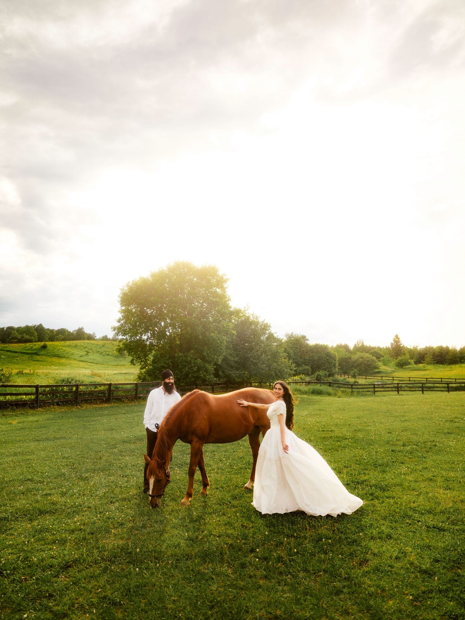 Bride in white gown and groom stand beside a chestnut horse on a lush green farm at golden hour.