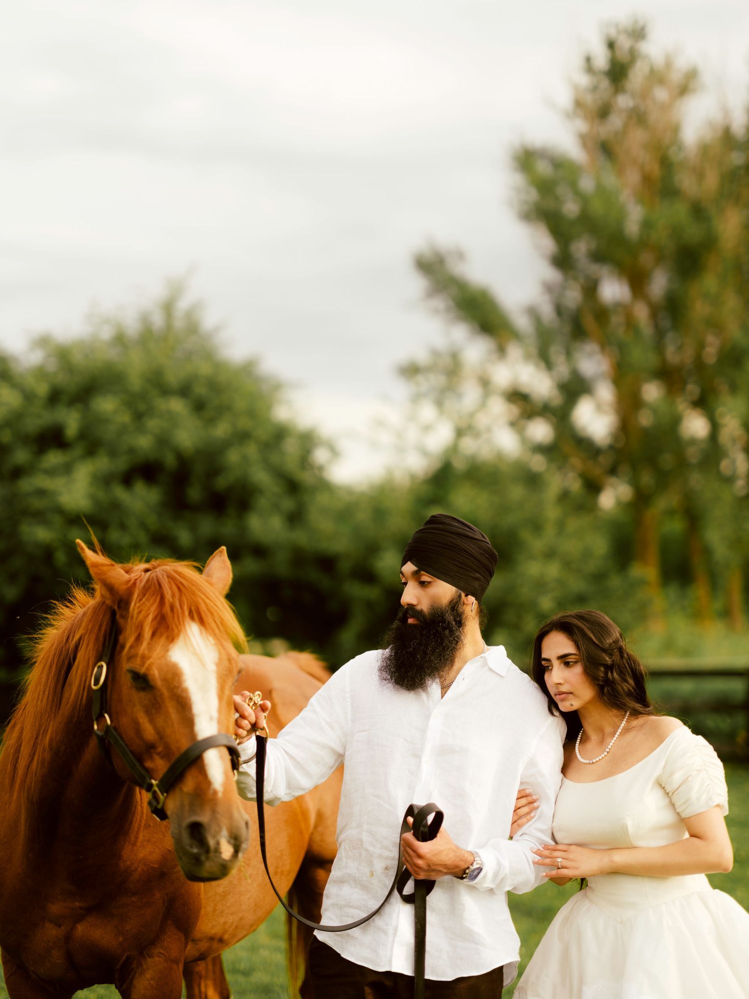 A bearded Sikh man in a turban and white shirt stands with a woman in a white dress beside a chestnut horse outdoors.