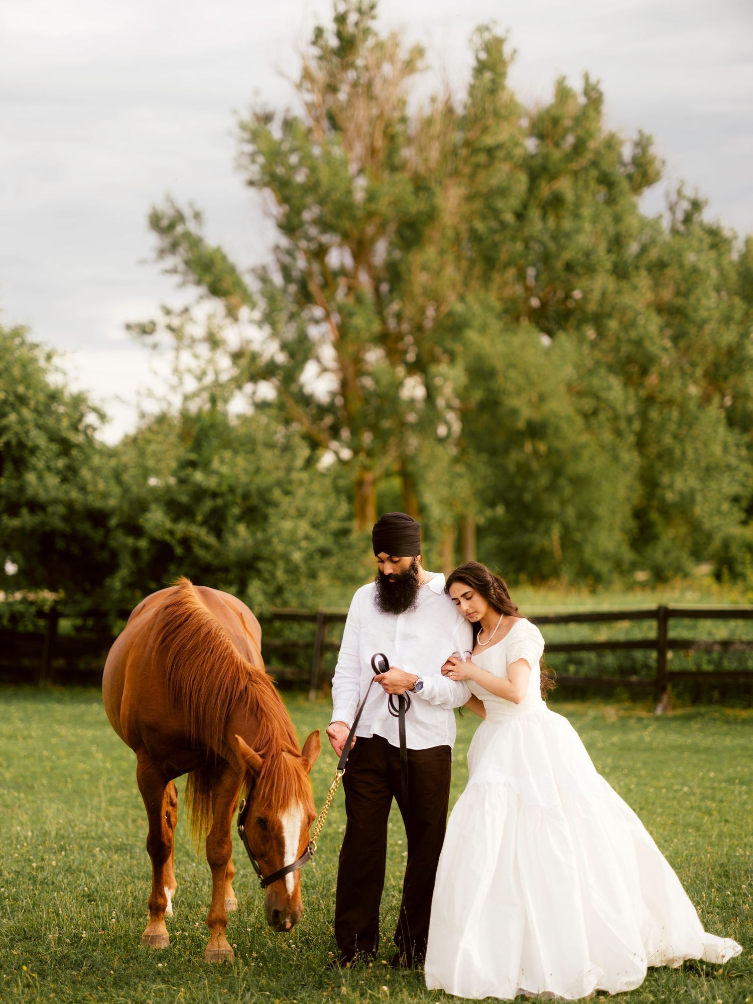Bearded groom in black turban and bride in white ball gown stand with a grazing chestnut horse outdoors.