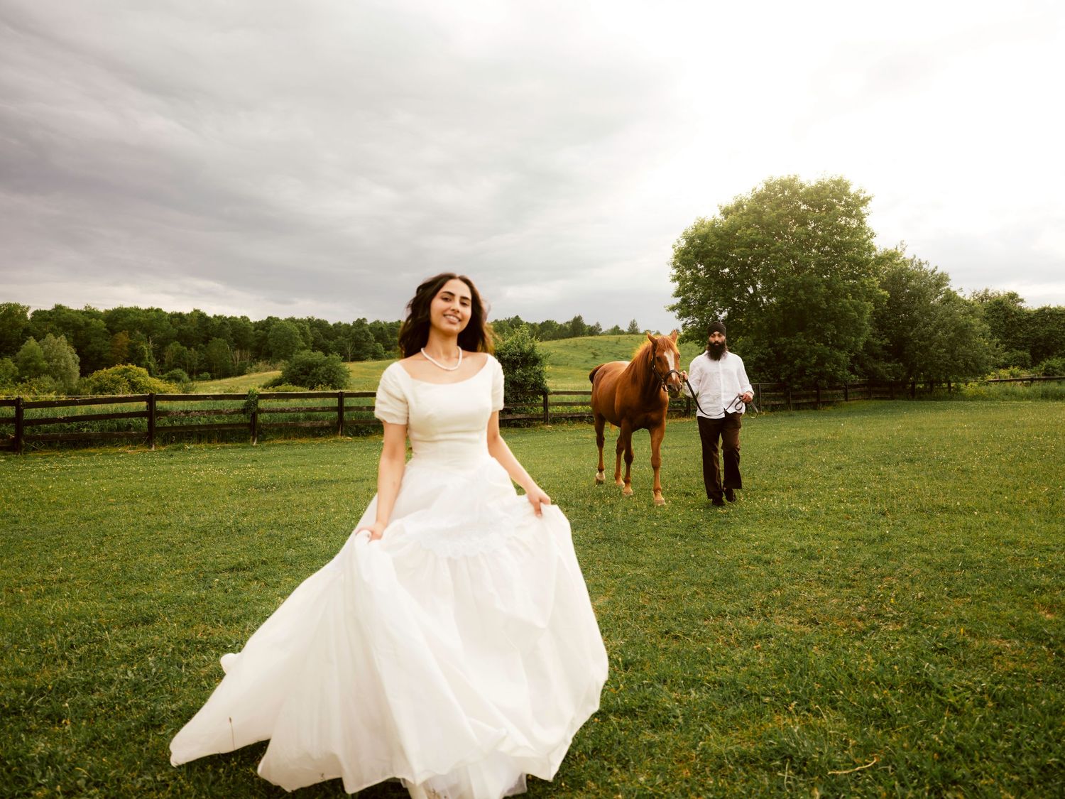 Bride twirls in white wedding gown on green farm field as groom walks with horse in background.
