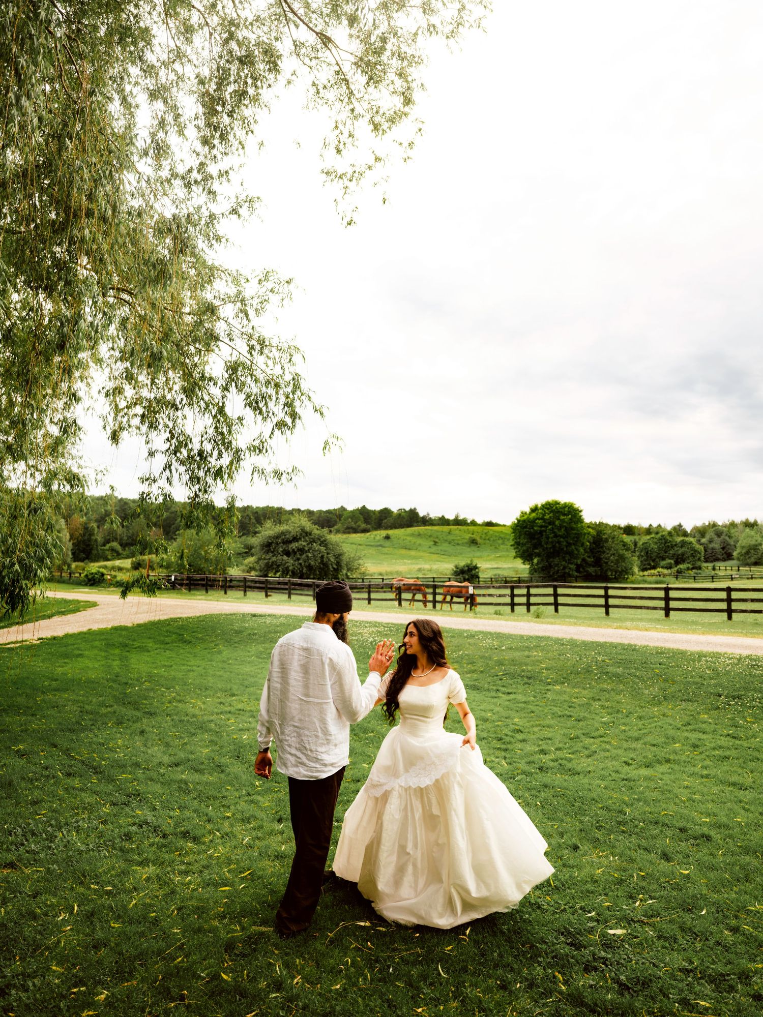Bride and groom dance on green lawn with a horse grazing in fenced pasture under cloudy skies.