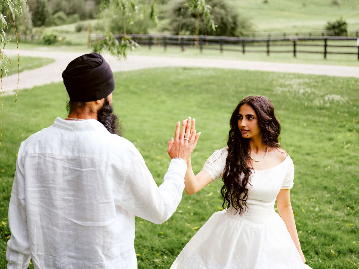 Bride and groom share a playful high-five moment outdoors near a wooden fence on a green farm.