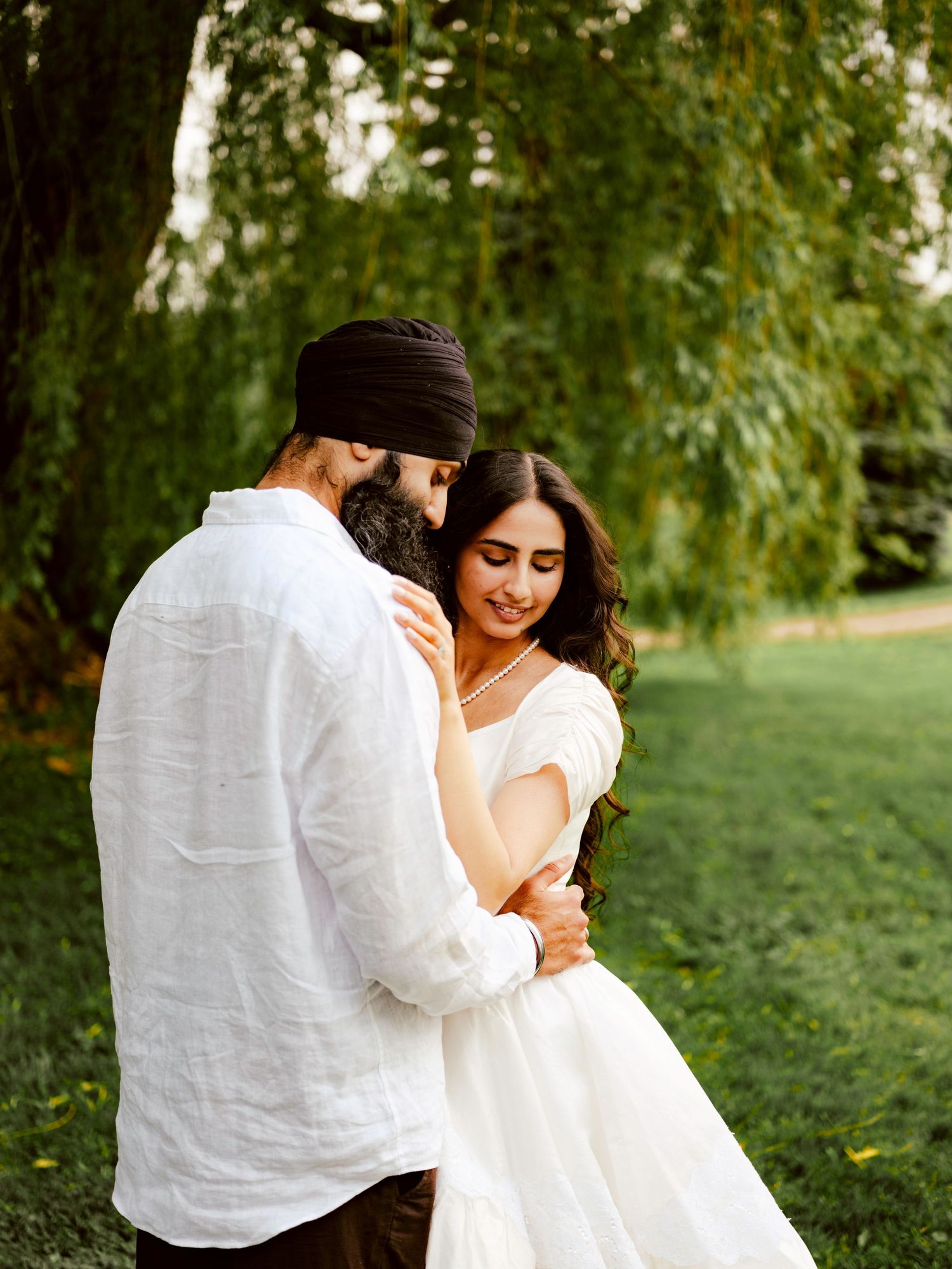 Sikh groom in black turban and white shirt embraces smiling bride in white dress under a weeping willow tree.