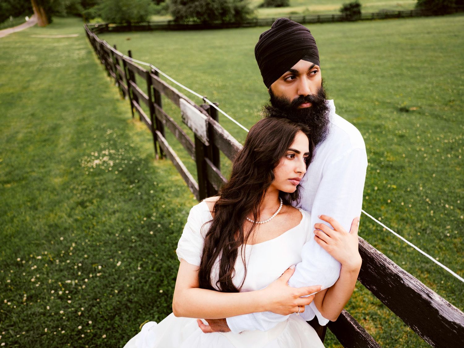 Sikh couple in white outfits share a tender embrace beside a wooden fence on a lush green farm.