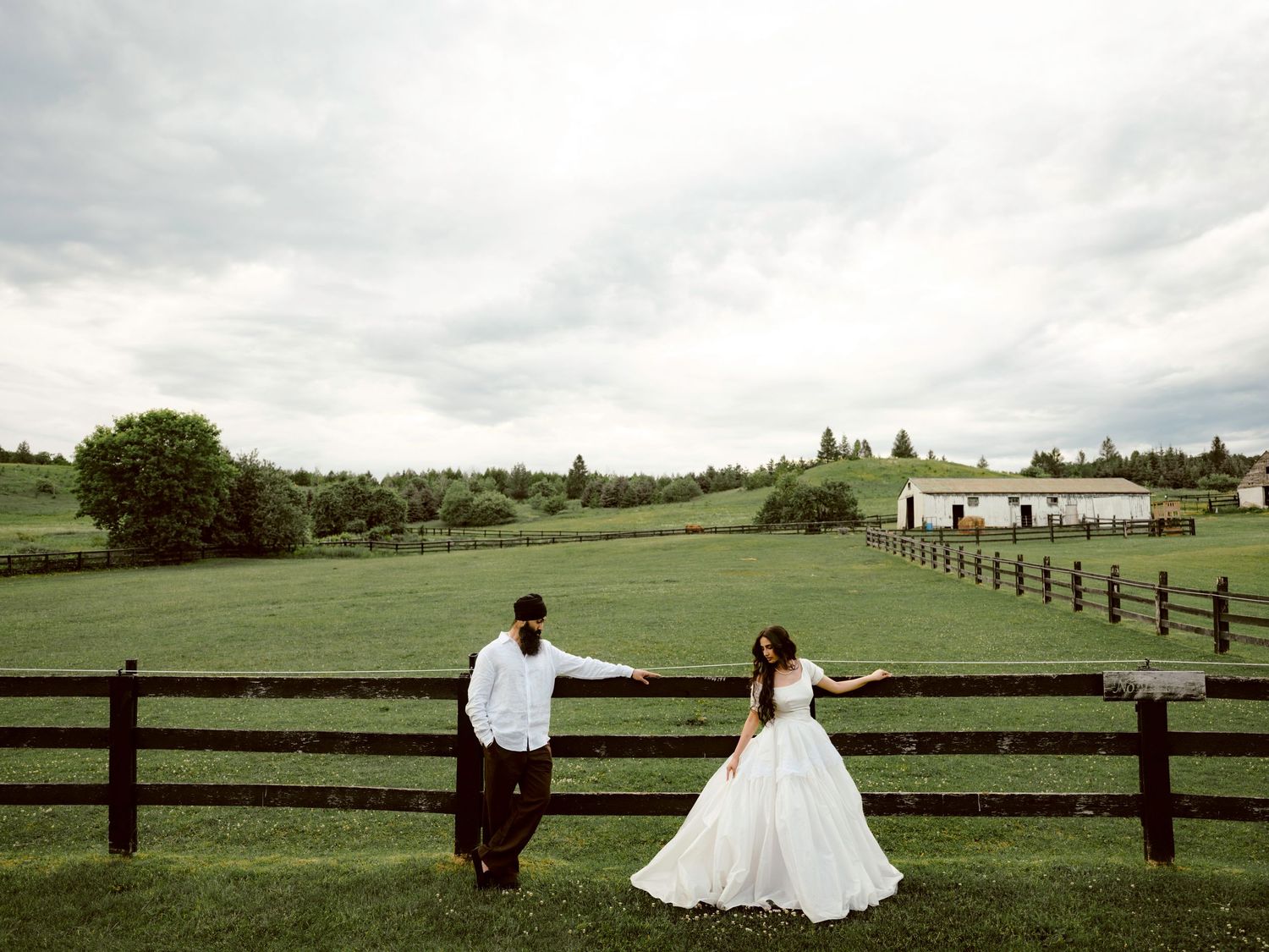Bride and groom holding hands by a rustic wooden fence on a lush green farm under cloudy skies.