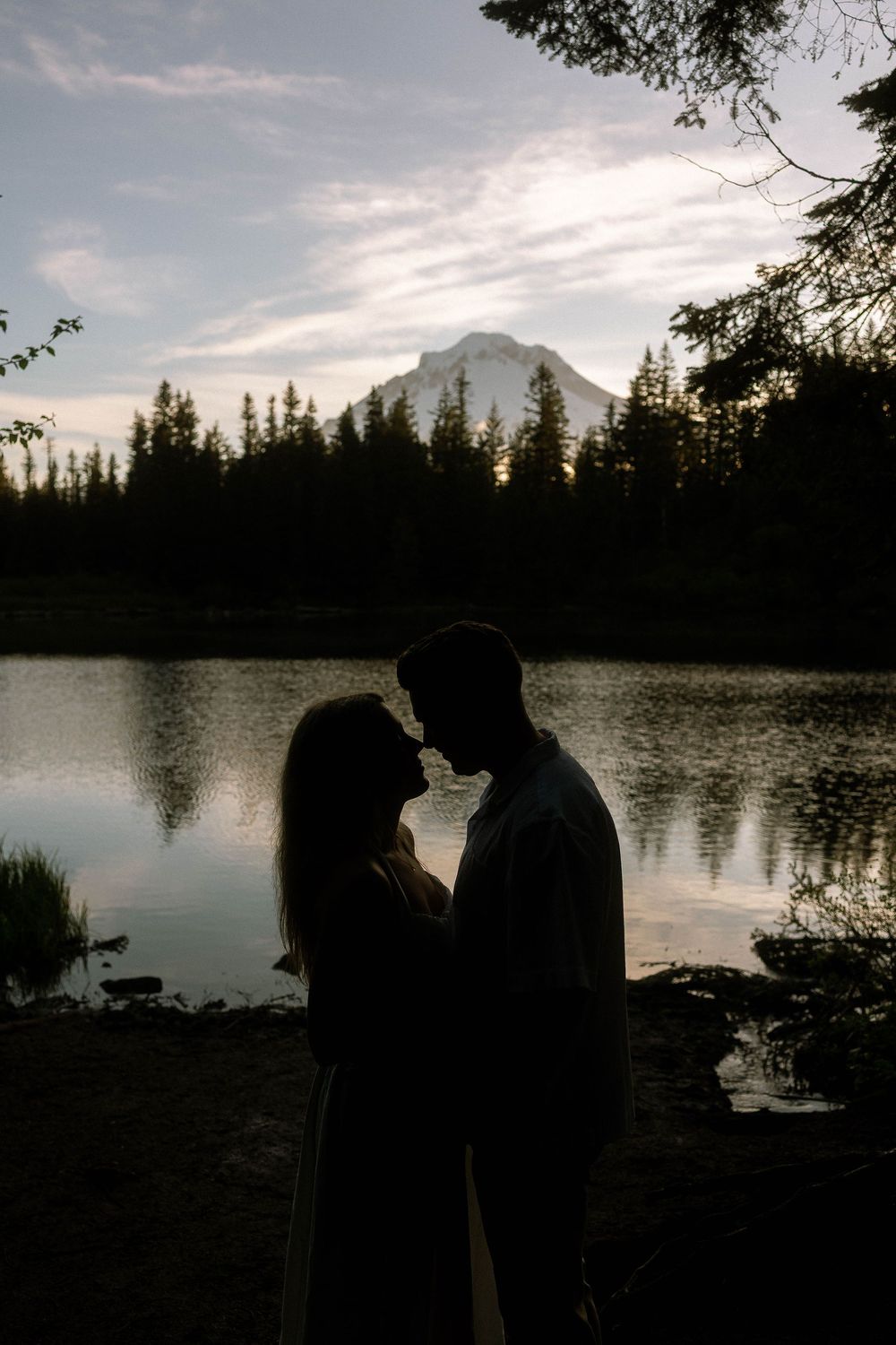 Silhouette of a romantic couple facing each other by a reflective mountain lake at dusk with evergreen trees.