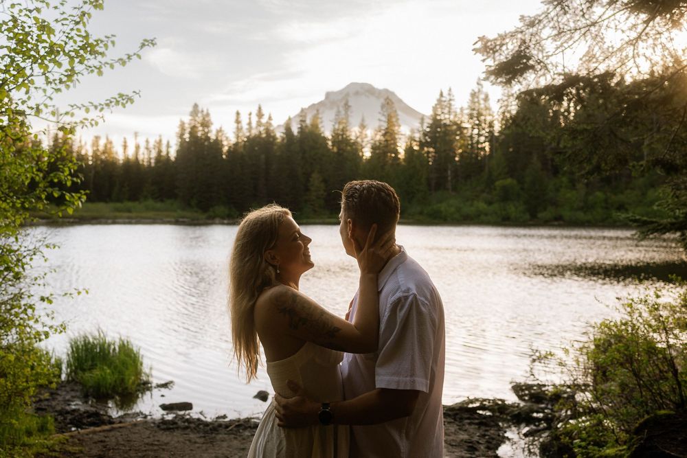 Couple embracing at golden hour by a mountain lake surrounded by pine trees with a peak in the background.