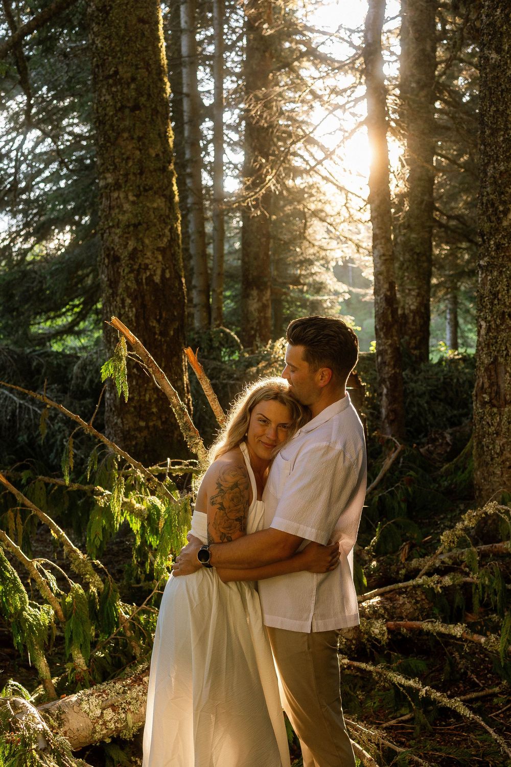 Couple embracing in sunlit forest, woman in white dress with tattoo, man in white shirt, golden hour light through tall trees.