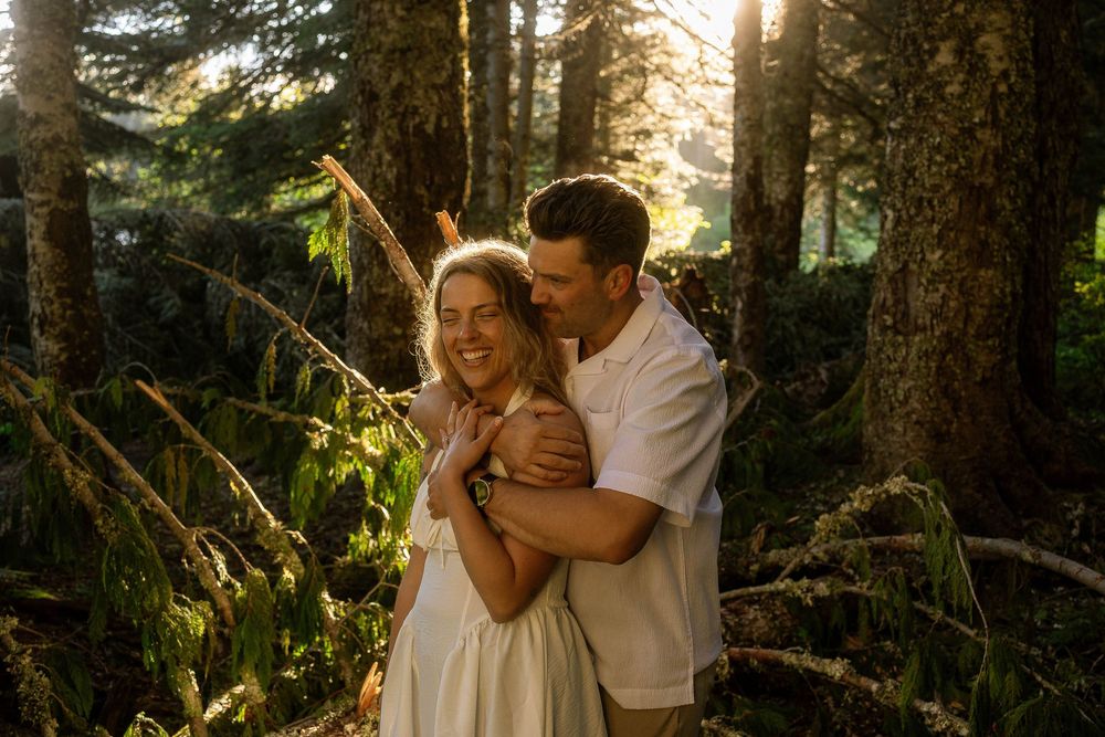 Couple embracing and laughing in sunlit forest with golden light filtering through tall pine trees.
