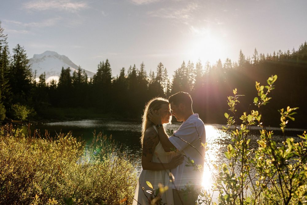 Couple embracing at golden hour by a mountain lake surrounded by evergreen trees with sunlight glowing behind them.