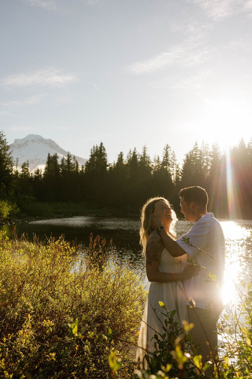 Couple embracing at golden hour by a mountain lake surrounded by evergreen trees with sun flare.